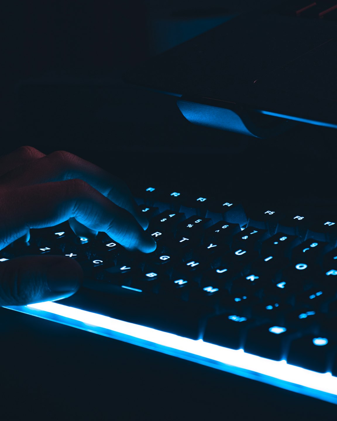 Tangan mengetik di keyboard mekanis dengan lampu latar dalam gelap.