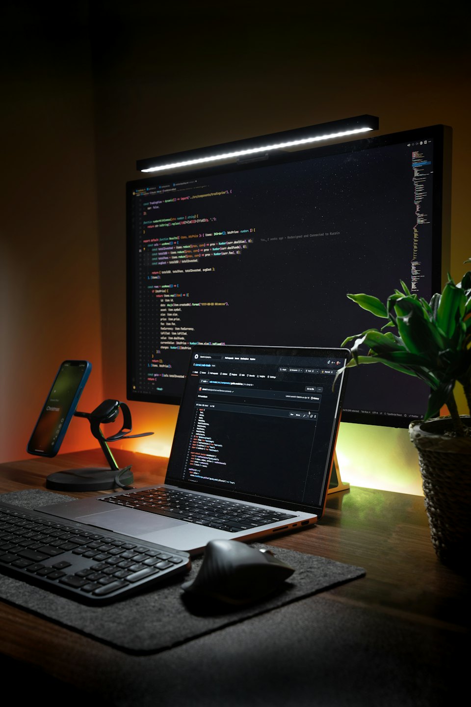 Meja pengembang dengan laptop, monitor, ponsel, dan tanaman.